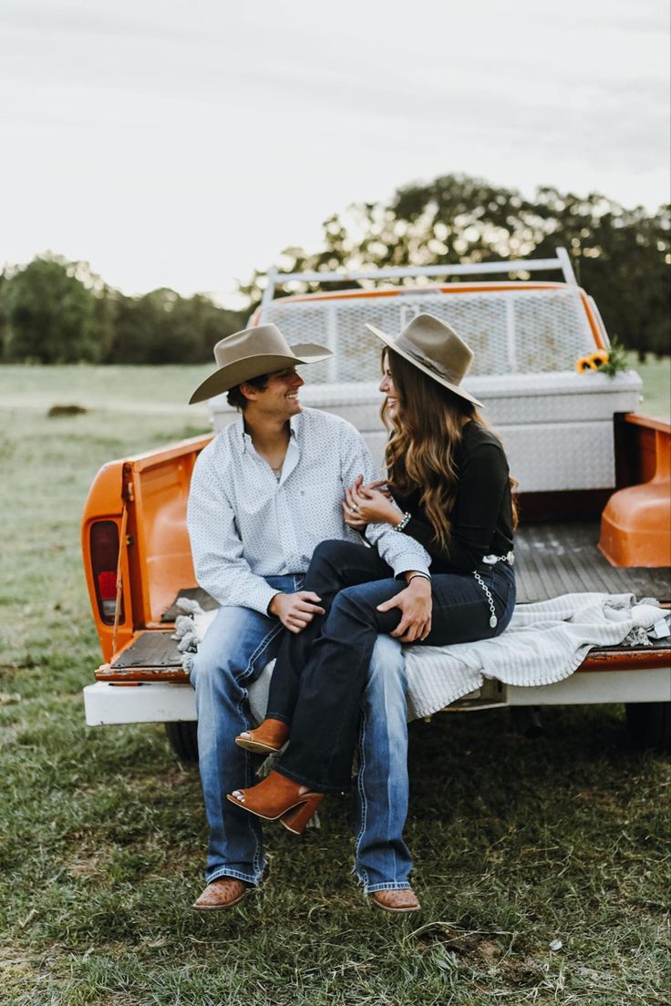 a man and woman sitting on the back of an orange pickup truck in a field