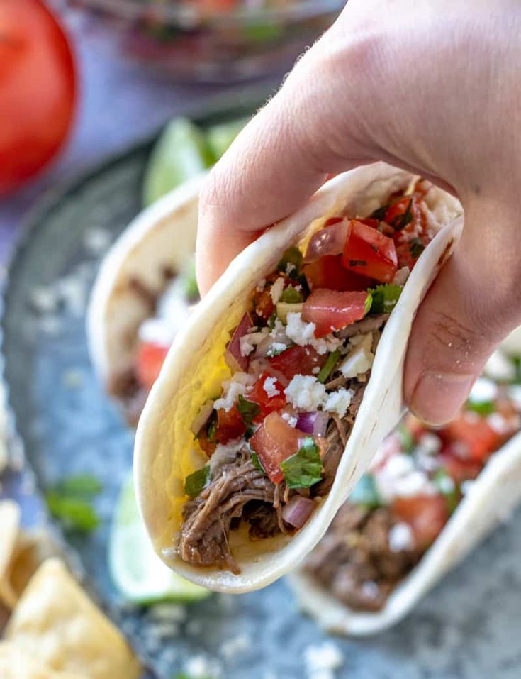 a hand holding a tortilla filled with meat, tomatoes and other toppings