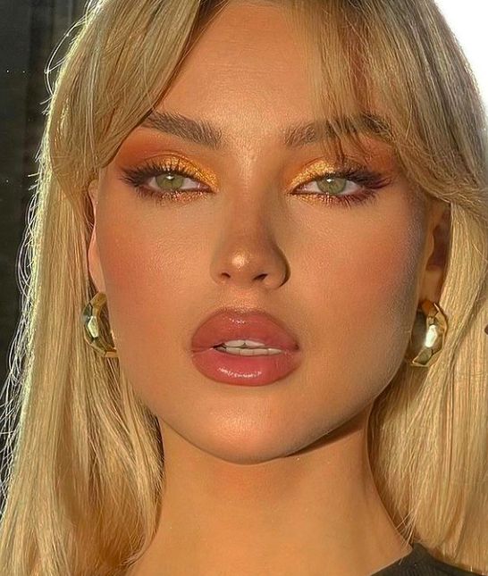 a woman with long blonde hair and bright makeup looks at the camera while wearing gold earrings