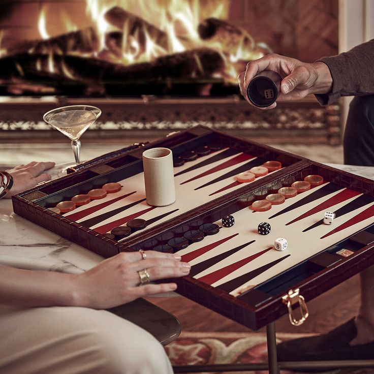 two people playing a game of backgamh in front of a fire place with drinks