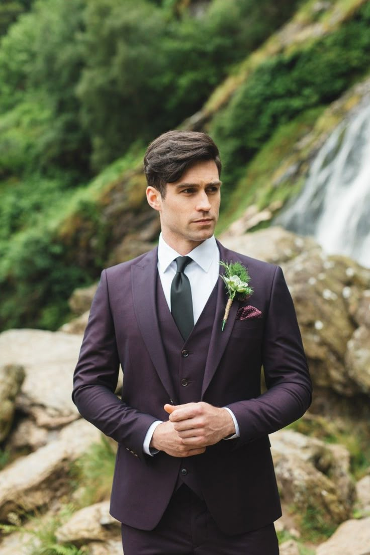 a man in a suit and tie standing next to a waterfall wearing a boutonniere