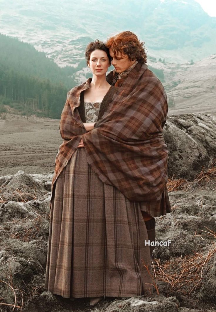 two women dressed in period clothing standing on a grassy hill with mountains in the background