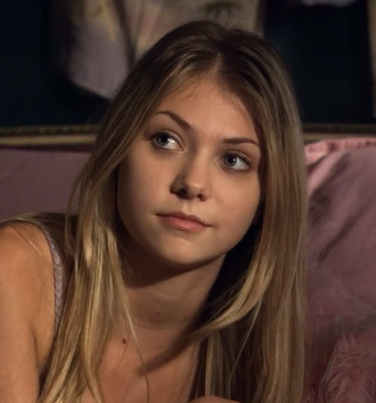 a young woman sitting on top of a bed next to a pink pillow and looking at the camera