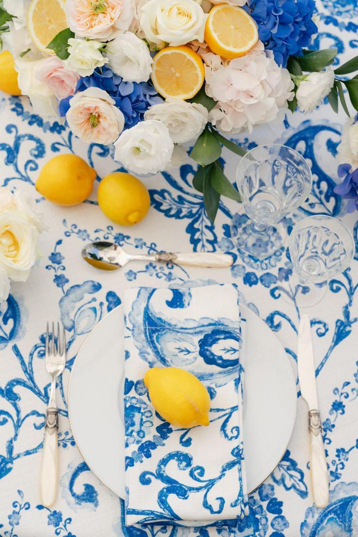 a blue and white table setting with lemons, flowers and silverware on it