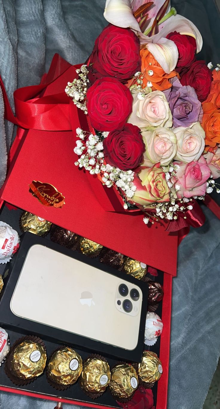 a red box filled with assorted chocolates next to a bouquet of roses and an iphone