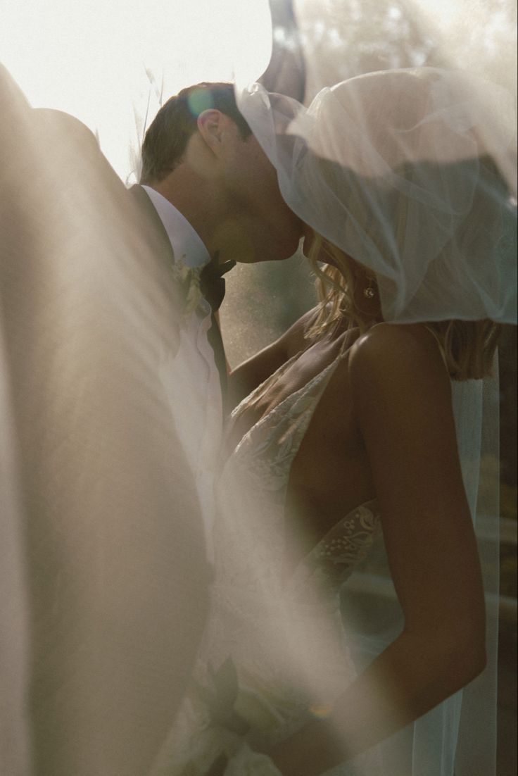 a bride and groom kissing in front of the sun with their veil blowing over them