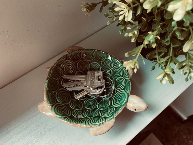 a green and white ceramic turtle with keys in it's mouth sitting on a shelf next to a potted plant