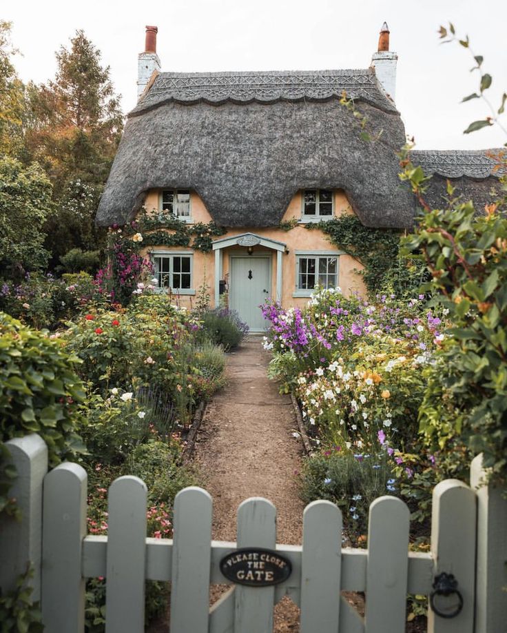 a house with a thatched roof surrounded by flowers and greenery in front of a white picket fence