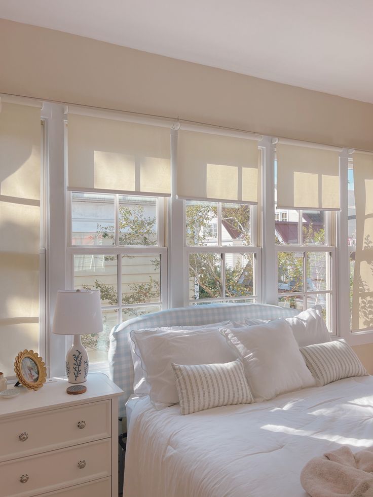 a white bed sitting in a bedroom next to two windows with shades on them and pillows