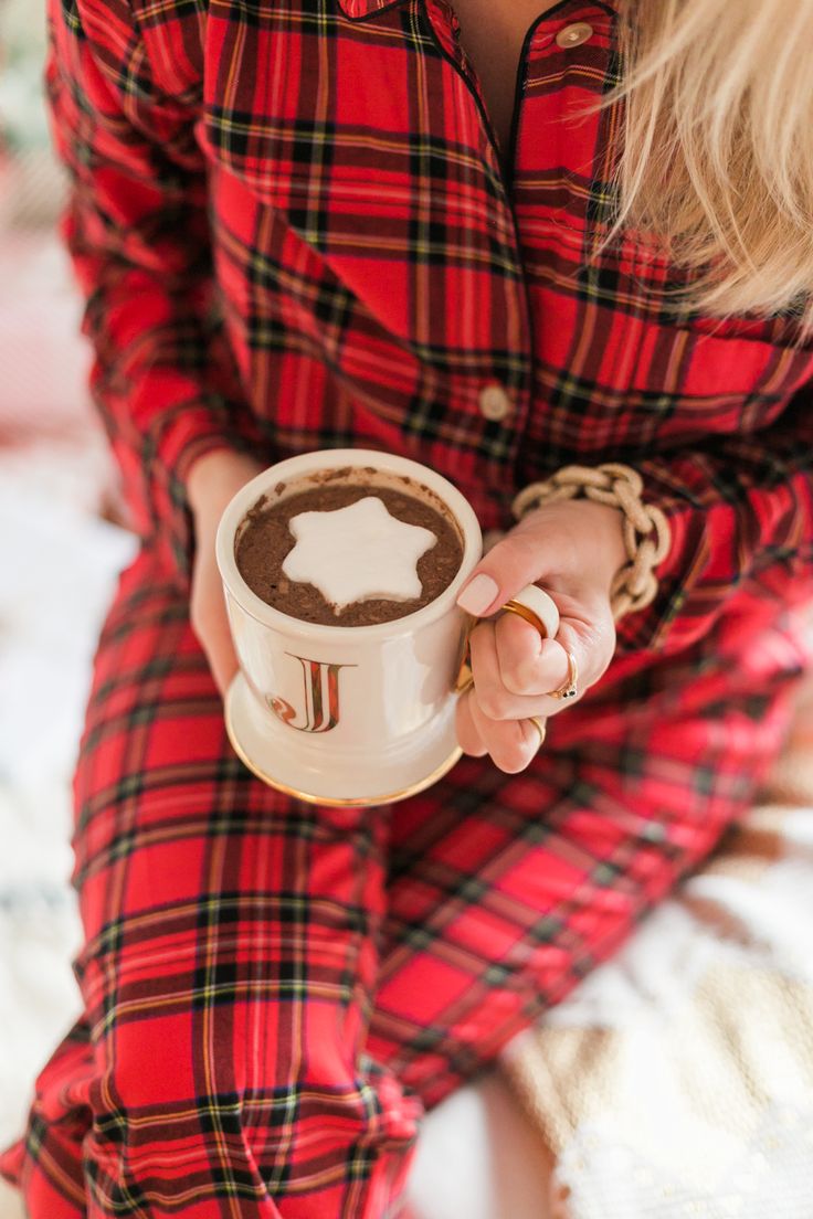 a woman in red pajamas holding a cup of hot chocolate on top of a bed