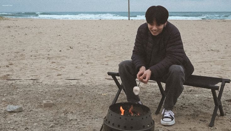 a man sitting on a bench next to an open fire pit with marshmallows in it