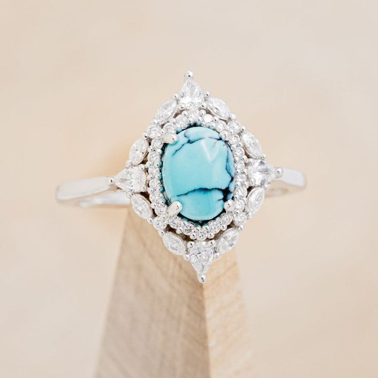 a ring with a turquoise stone surrounded by white diamonds on top of a wooden stand