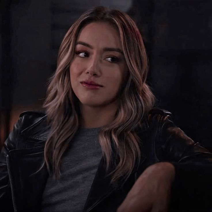 a woman with long hair wearing a black leather jacket and looking at the camera while sitting down