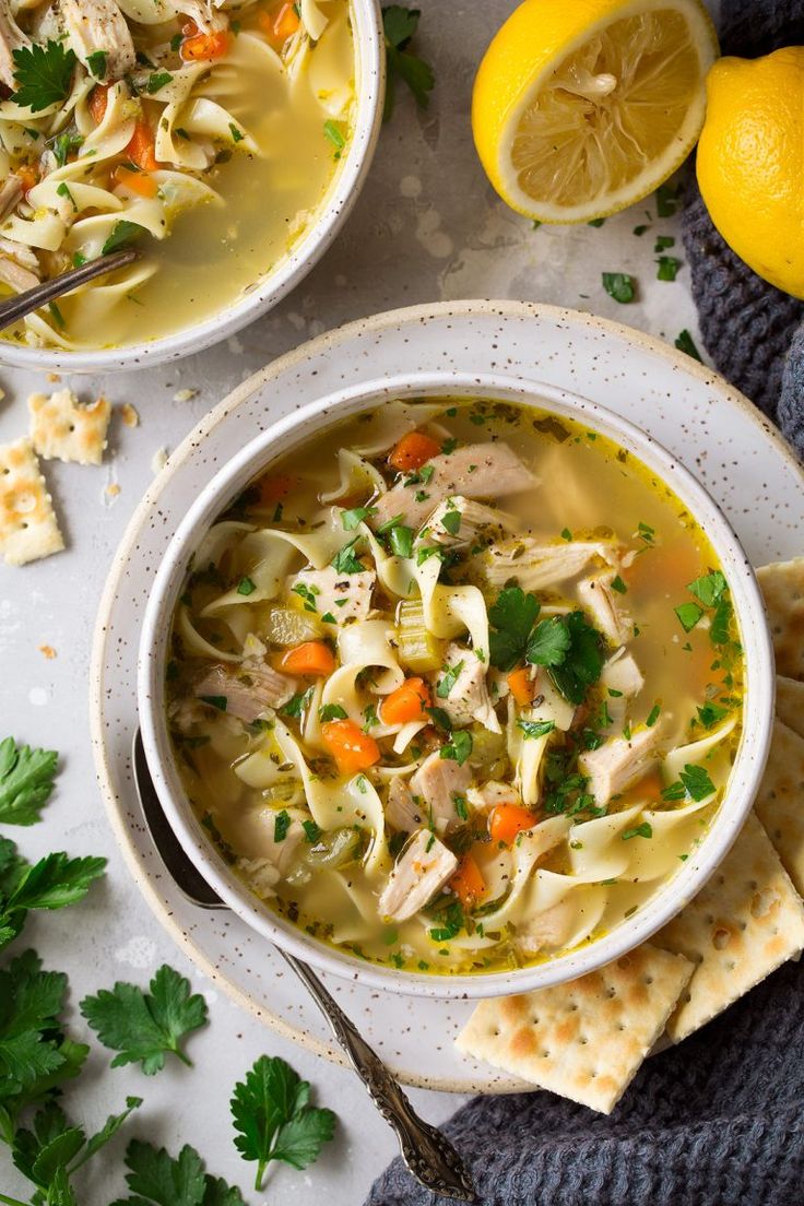 two bowls filled with chicken noodle soup next to crackers and lemon wedges