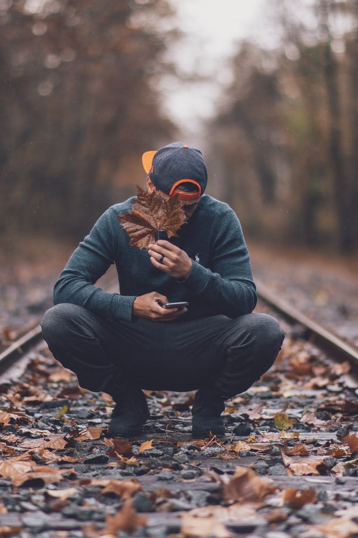 a man squatting down on the train tracks looking at his cell phone while holding an autumn leaf