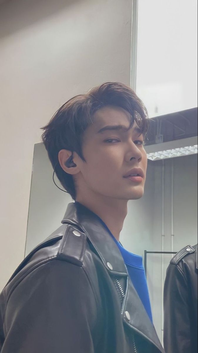 a young man in a black leather jacket looking at his earphones while standing in front of a mirror