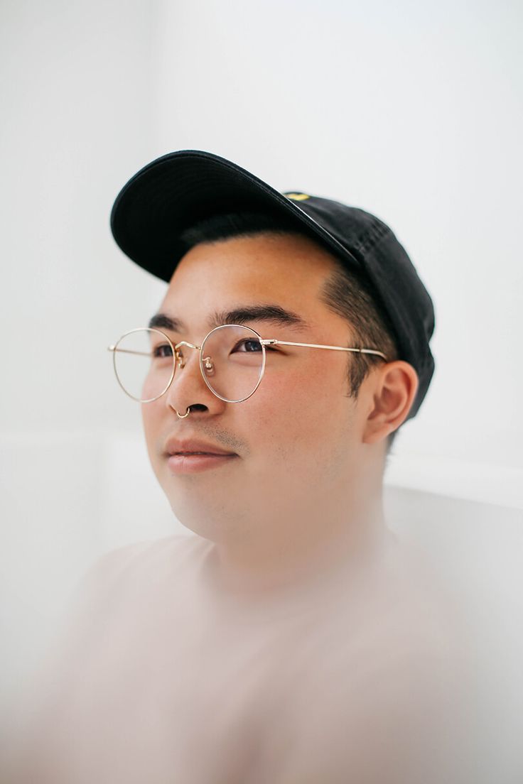 a man wearing glasses and a baseball cap looks off into the distance while standing in front of a white wall