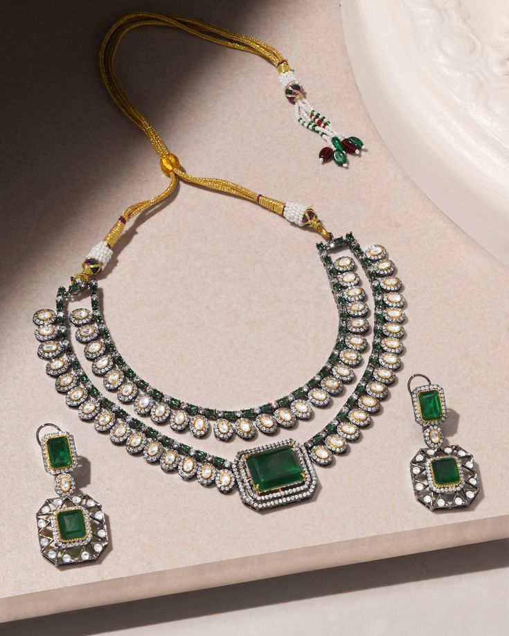 Presenting a captivating two-layer Polki set featuring a stunning emerald Polki stone at its center, accompanied by matching earrings. This ensemble exudes drama and elegance, perfect for adding a touch of regal charm to any occasion. Finish: Rhodium Material: Brass, Polkis & Emerald Stones Color: White, Green Size: Free Size, Adjustable Closure Type: Draw String Box Contains: 1 Necklace, 1 Pair of Earrings Elegant Emerald Kundan Necklace With Stone Work, Elegant Kundan Necklace With Emerald Stone Work, Elegant Emerald Kundan Necklace For Festive Occasions, Elegant Kundan Emerald Necklace For Festive Occasions, Hand-set Emerald Necklace For Celebration, Traditional Emerald Kundan Necklace For Formal Occasions, Elegant Green Kundan Necklace For Festive Occasions, Festive Hand Set Emerald Necklace, Festive Green Jewelry Sets With Elegant Design