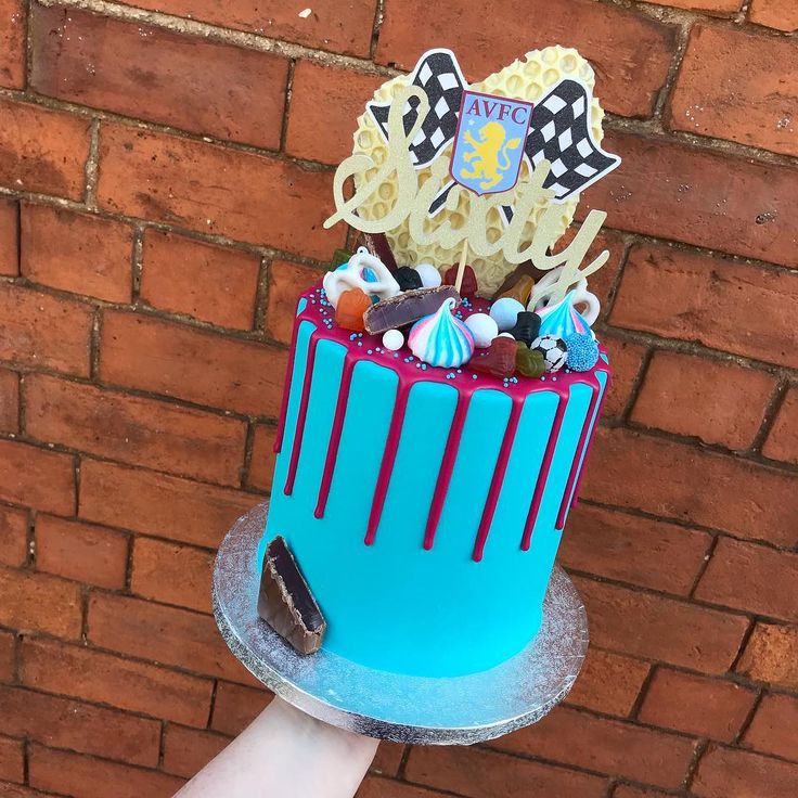 a hand holding a blue cake with red icing and decorations on top, against a brick wall