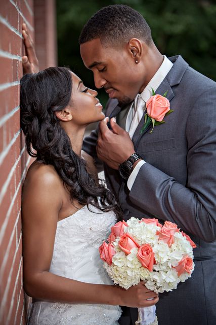 a bride and groom standing next to a brick wall with flowers in their hands, looking into each others eyes