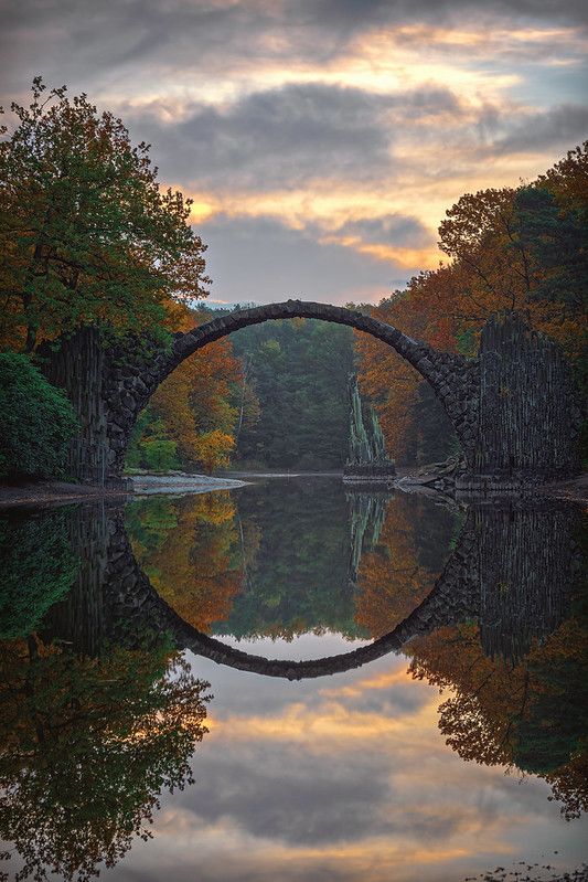 a bridge that is over some water with trees in the background and clouds in the sky