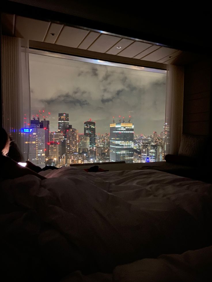 a person sitting on a bed in front of a window looking out at the city