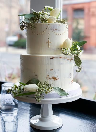 a three tiered cake with flowers and greenery on the top is sitting on a table in front of a window