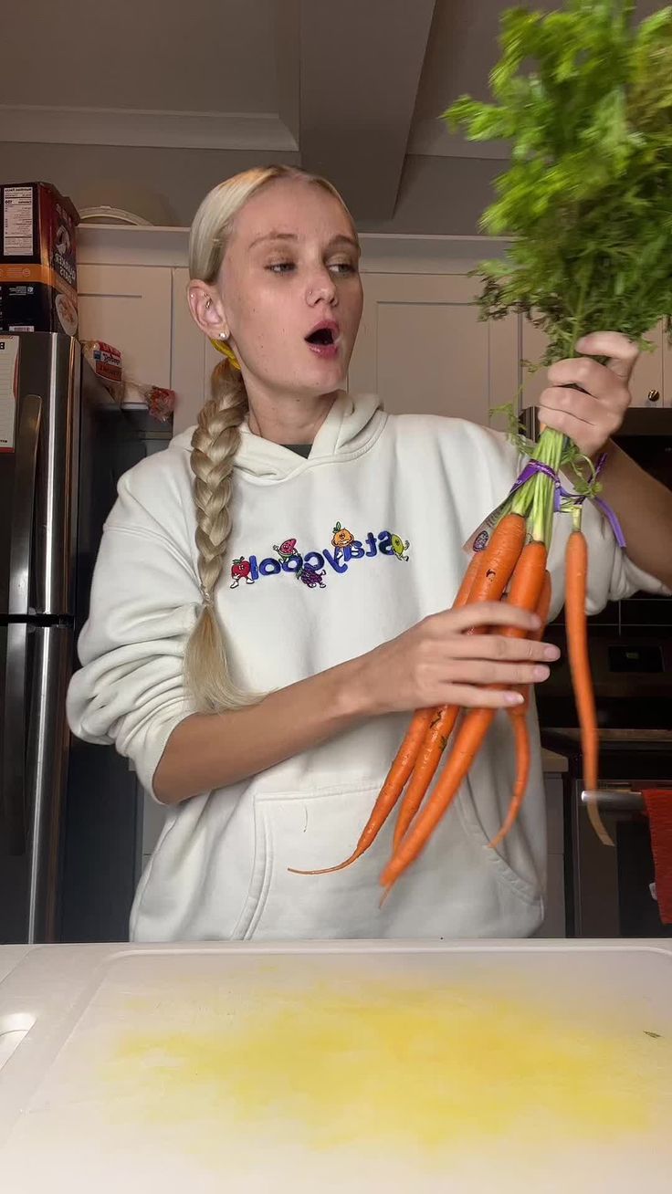 a woman holding two carrots in front of her face while standing next to a counter