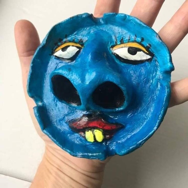 a hand is holding up a blue clay face with yellow eyes and mouth painted on it