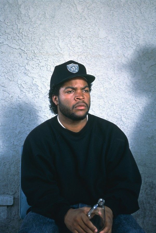 a man with a black hat is sitting in front of a wall and holding a bottle