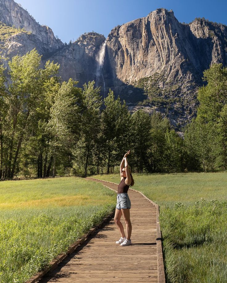 a woman is standing on a wooden walkway in front of mountains and trees, reaching up into the sky