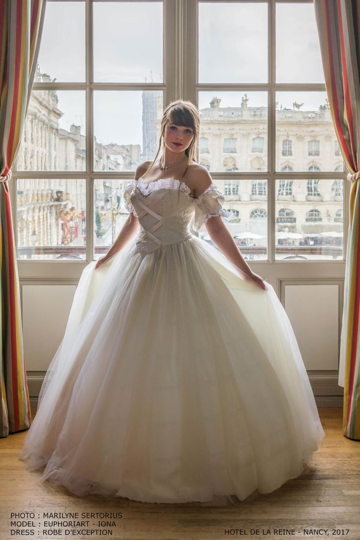 a woman in a ball gown standing by a window with her hands on her hips