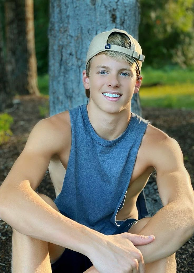 a young man sitting on the ground in front of a tree wearing a hat and blue shirt