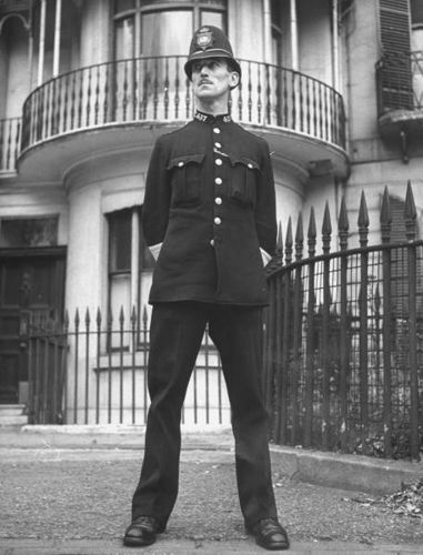 a black and white photo of a man in uniform standing on the sidewalk near a building