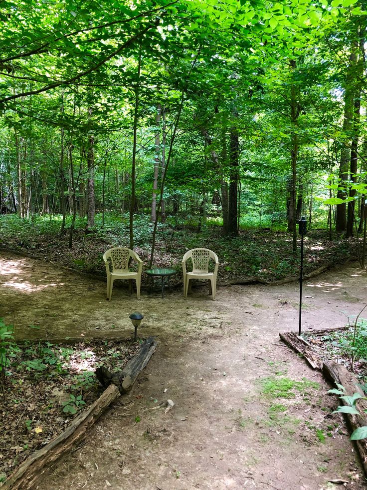 two white chairs sitting on top of a dirt road in the middle of a forest