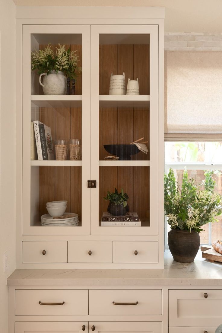 a white china cabinet with glass doors and drawers in a kitchen next to a window