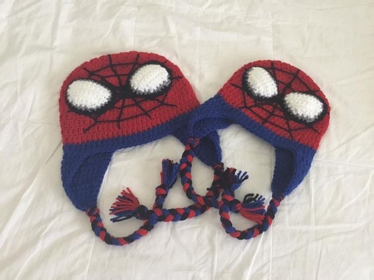 two crocheted hats with spiderman faces on them sitting on a white sheet