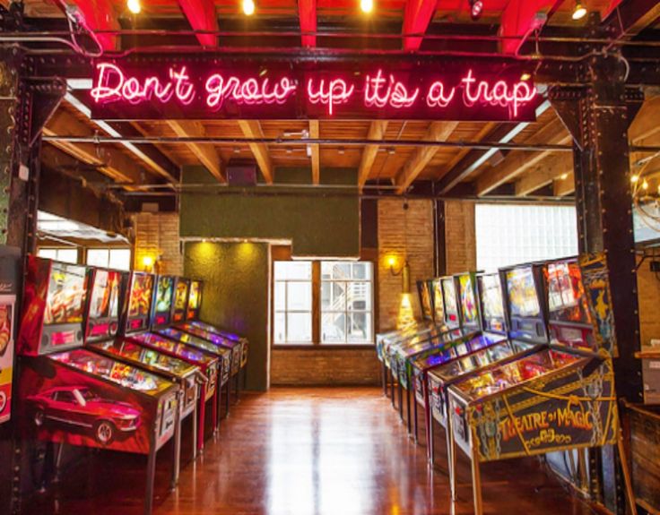 an arcade with pinball machines and neon signs on the ceiling that says don't grow up it's a trap