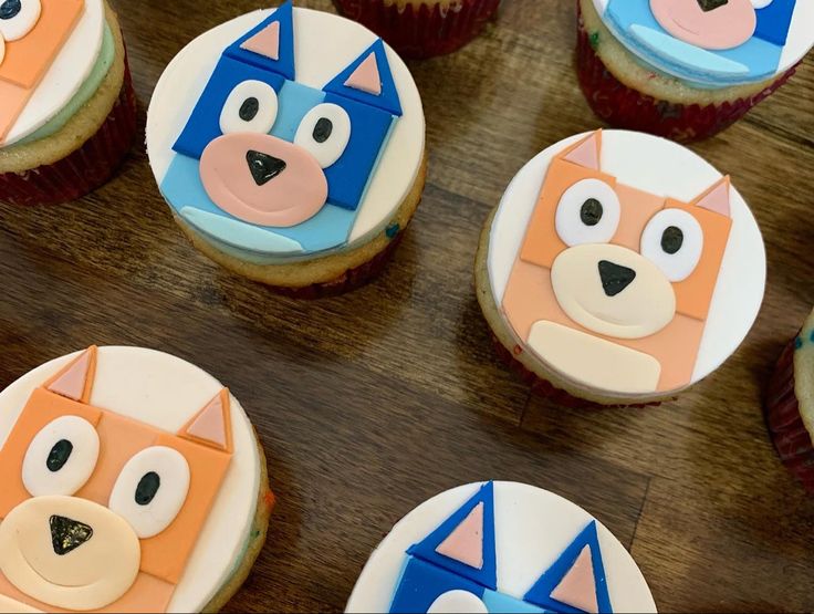 cupcakes decorated like cartoon cats and dogs on a wooden table with other cupcakes in the background