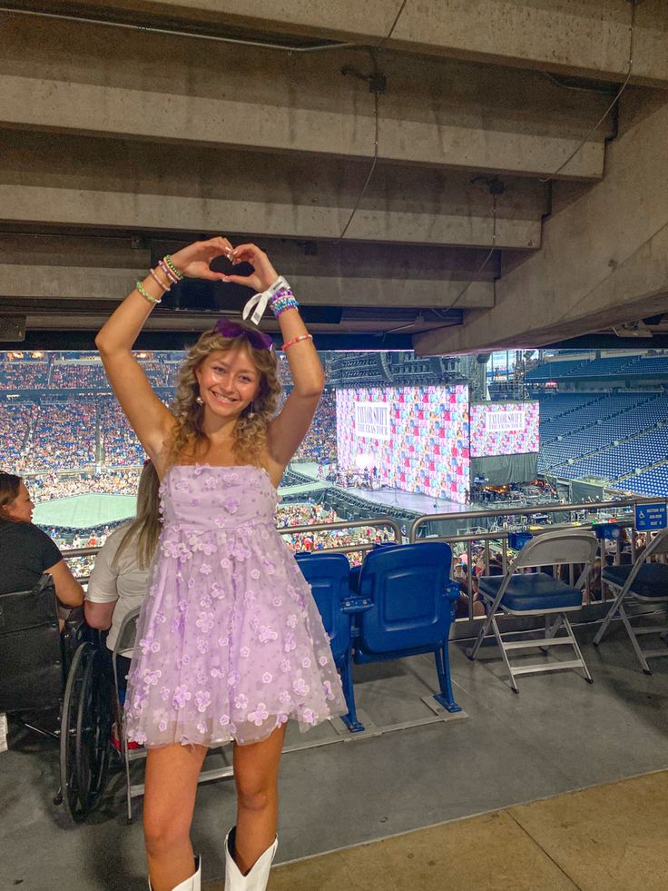 a woman in a purple dress and cowboy boots at a baseball game holding her hands up above her head