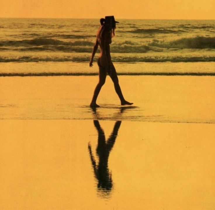 a woman walking on the beach with her reflection in the wet sand and water behind her