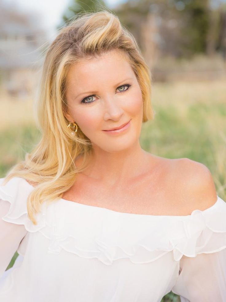 a woman with blonde hair and blue eyes wearing a white top in a field smiling at the camera