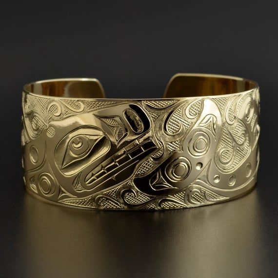 This cuff is hand-engraved and measures 1" wide by 6" long and was designed by First Nations artist Kelvin Thompson. This will fit a wrist between 6 1/4" and 6 1/2" in circumference. Custom lengths can be made on request. This piece is made to order and is titled 'Pod of Whales'.Whales usually signify longevity and strong community bonds due to their tendency to travel in close multi-family pods. In artworks, Orcas can be identified by a large, snub-nosed head; elongated nostrils; a wide, toothy Luxury Engraved Cuff Bracelet For Anniversary, Unique Yellow Gold Cuff Bracelet For Formal Occasions, Luxury 14k Gold Etched Jewelry, Ceremonial Gold Bracelets With Polished Finish, Symbolic Carved Jewelry For Formal Occasions, Luxury Engraved Bracelets For Ceremonial Occasions, Engraved Gold Cuff Bracelet For Formal Events, Luxury Engraved Gold Bracelet For Ceremonial Occasions, Engraved Gold Cuff Bracelet For Formal Occasions