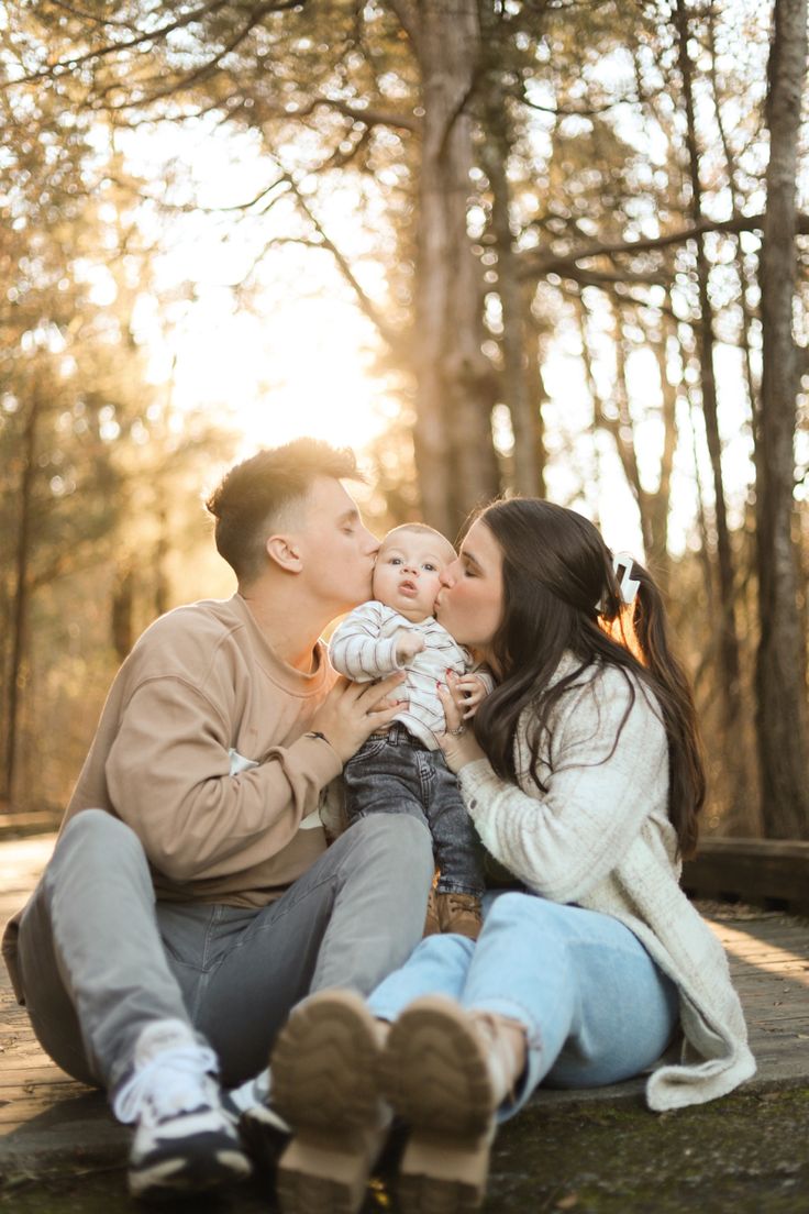 a man and woman kissing while holding a baby in their lap, with the sun shining through the trees behind them