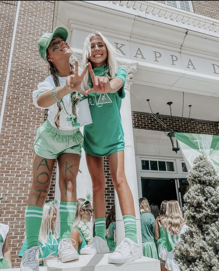 two women dressed in green and white standing on top of a building with their hands up