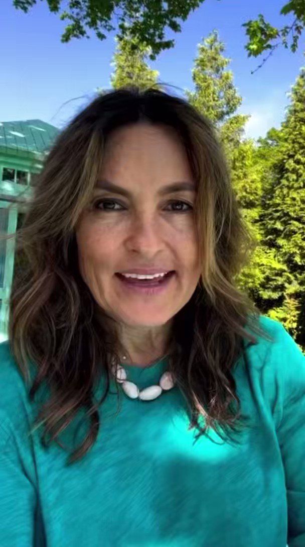 a woman with long hair wearing a blue shirt and smiling at the camera while standing in front of some trees