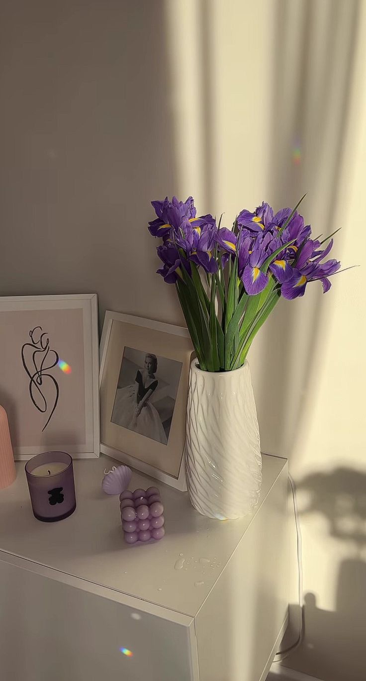 a white vase filled with purple flowers sitting on top of a table next to pictures