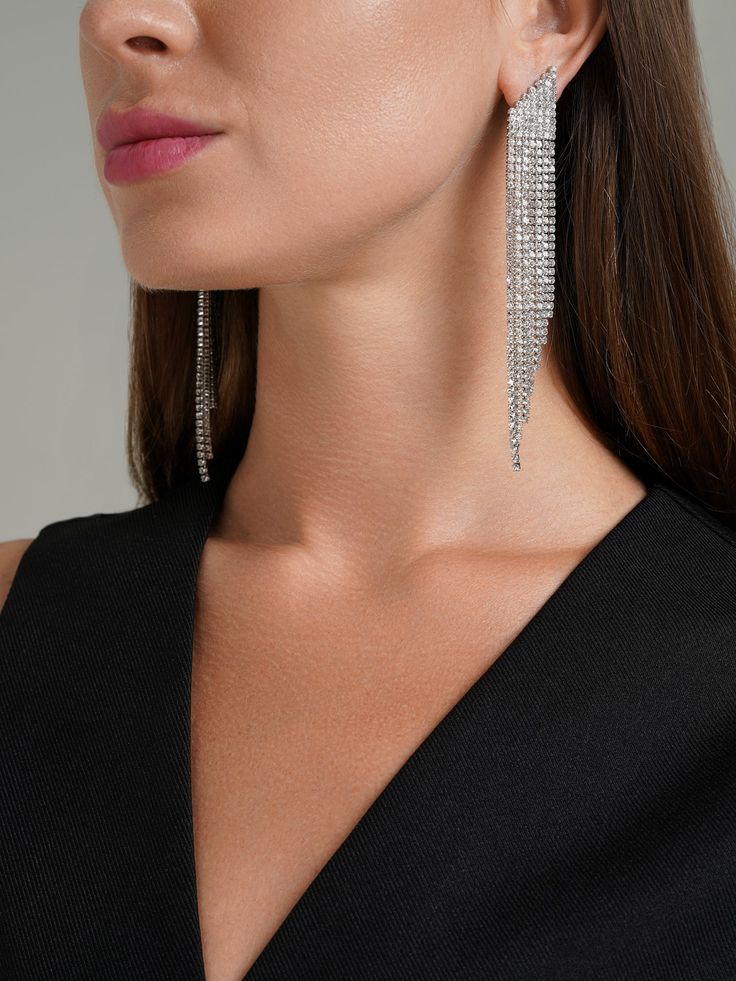 These earrings are striking, silver, and feature a cascading design. They are embellished with multiple rows of sparkling crystals, creating a dazzling effect that catches the light beautifully. The earrings taper towards the ends, giving them a sleek, elegant appearance. Perfect for adding a touch of glamour to any outfit, these earrings are ideal for special occasions or when you want to make a statement. Glamorous Silver Sparkling Crystal Earrings, Party Jewelry With Rhinestones In Long Drop Shape, Silver Rhinestone Chandelier Earrings For Evening, Party Jewelry With Long Drop Rhinestones, Long Drop Rhinestone Party Jewelry, Party Long Drop Rhinestone Jewelry, Silver Crystal Earrings With Rhinestone Fringe, Silver Chandelier Earrings With Bling For Evening, Silver Bling Chandelier Earrings For Evening