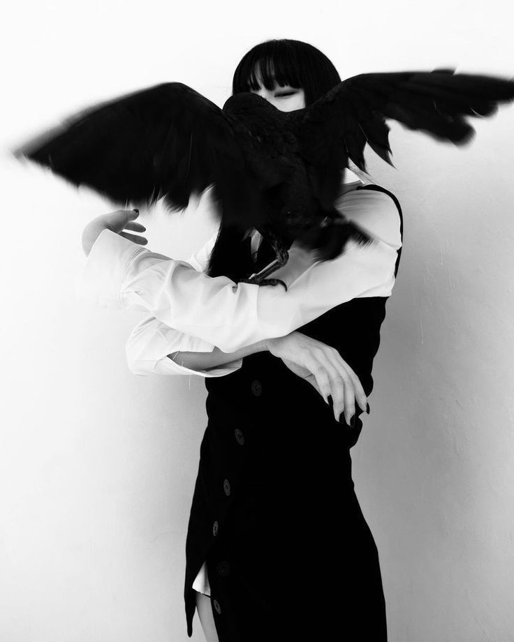 a woman holding a black bird in her arms while standing next to a white wall