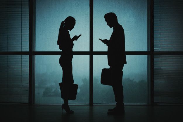 two people standing in front of a window looking at their cell phones